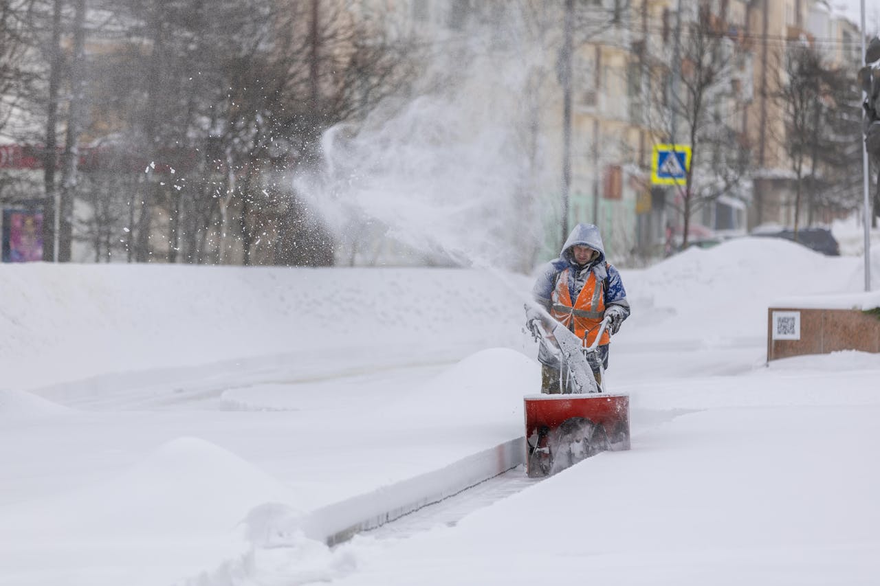 About Person using a snow blower to clear a city sidewalk during a heavy winter snowfall.