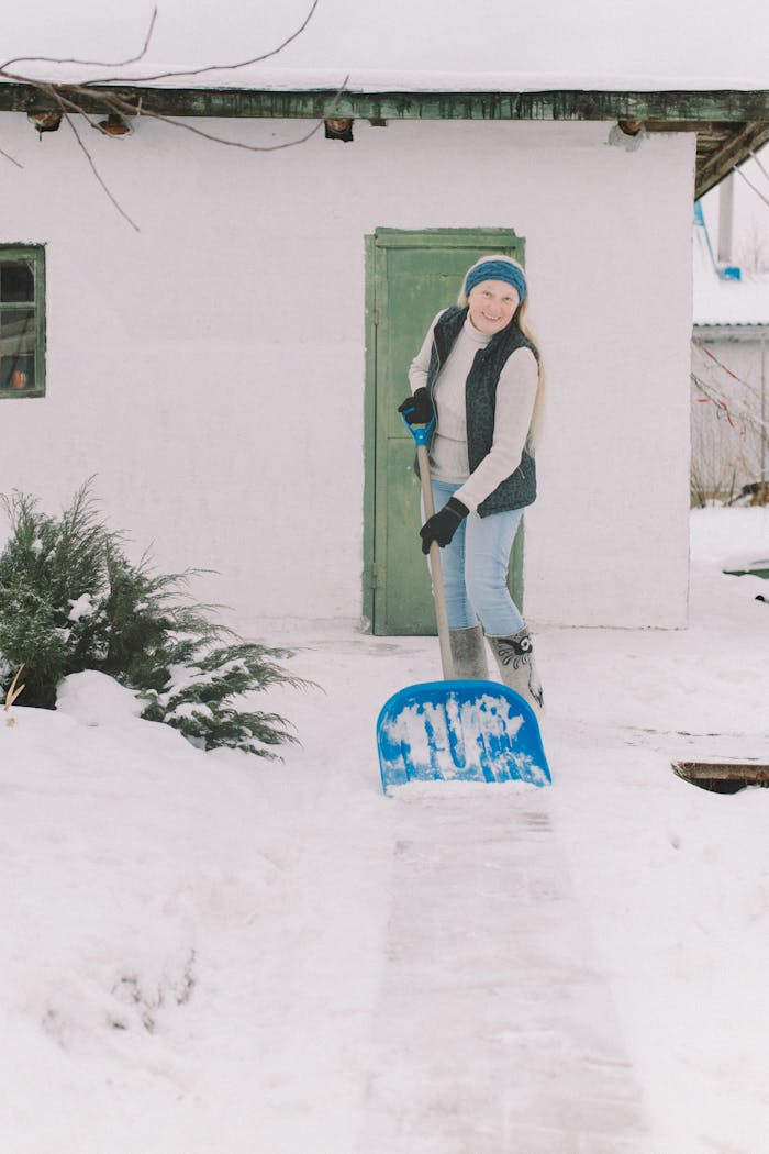 Services Smiling woman shoveling snow in a backyard with a white shed on a winter day.