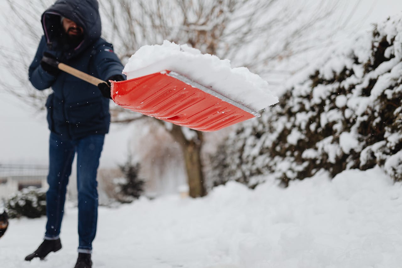 About A man uses a red shovel to clear snow in a snowy yard, emphasizing winter maintenance.